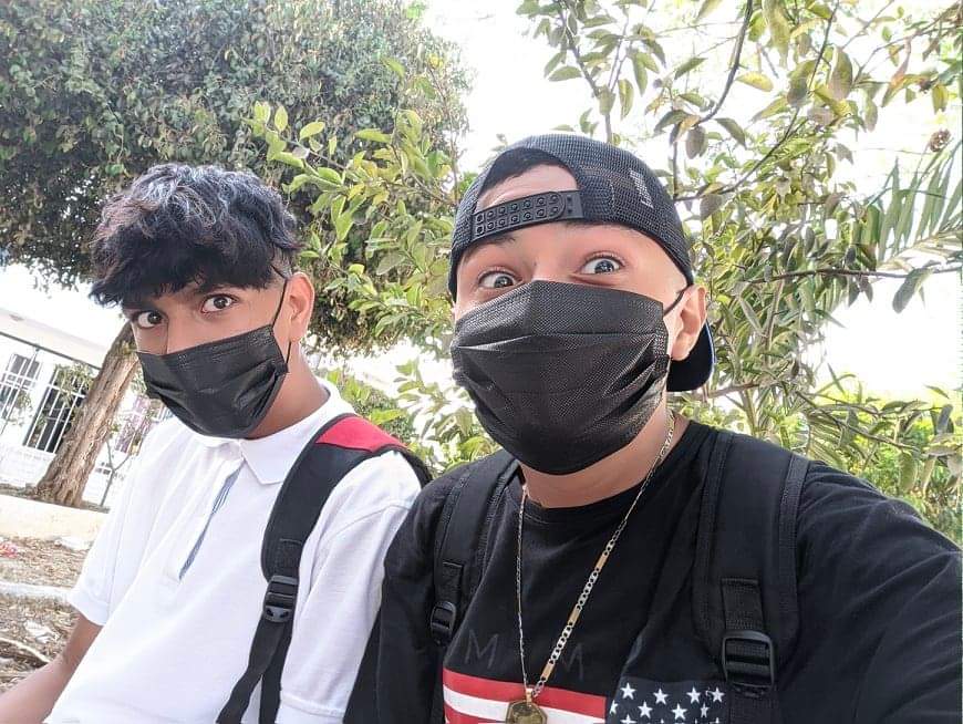 JX y Axael sentados bajo los árboles, con tapabocas negros, mirando a la cámara con cara de estar pasándola bien.